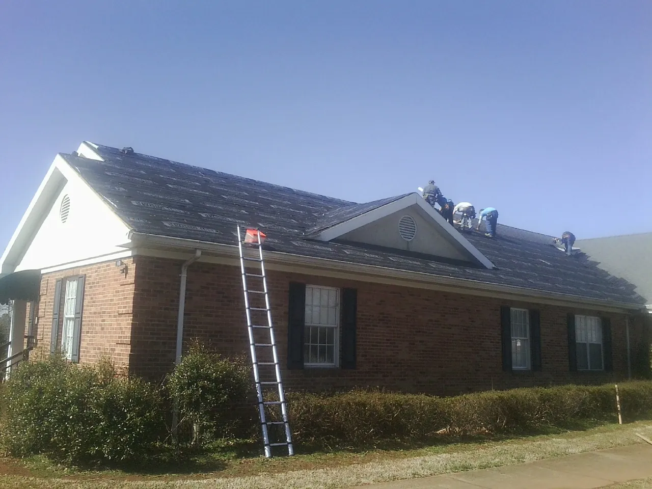 Crew tearing shingles off a brick ranch roof with a ladder set — Metal Roof Repair work by our crew in Ortonville