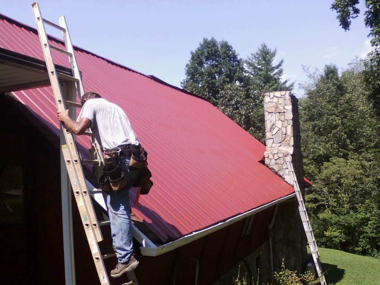 Roofer on a ladder beside a red metal roof and stone chimney — Soffit & Fascia Repair by Roofing Brothers in Ortonville